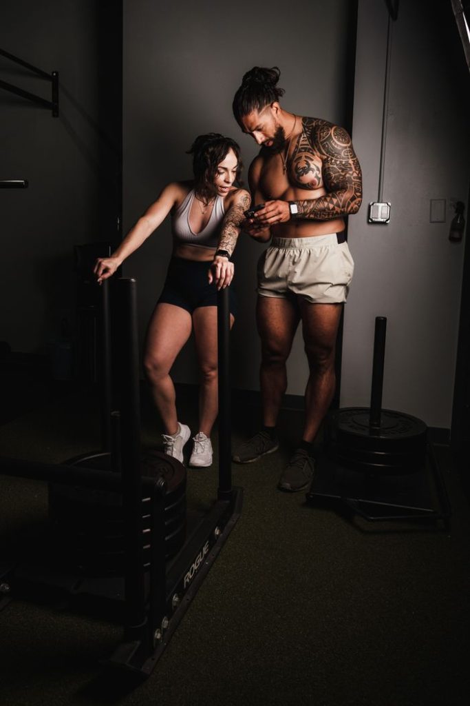 A tattooed couple stands in a gym, checking a smartphone during a fitness session.